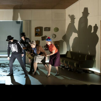 In a large room with one wall open toward the audience and at least 15 clocks on the back wall, a group of five actors leans over to look at something, with stark shadows cast on the wall behind them.