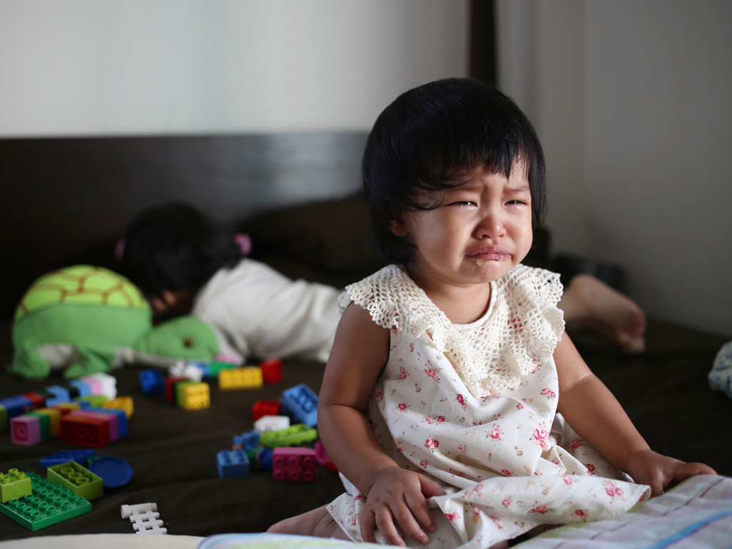 crying little girl surrounded by toys sitting on bed