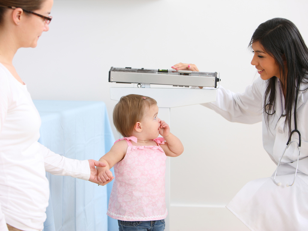 A healthcare professional weighing a toddler while their parent holds their hand