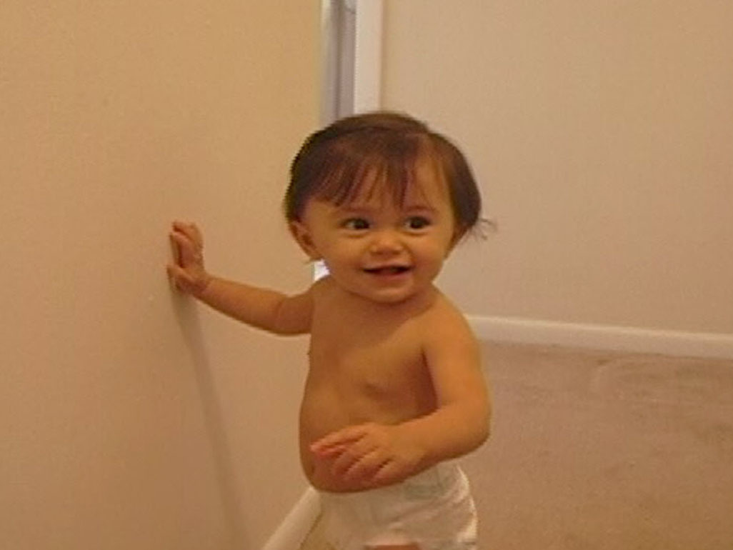 A baby in diapers standing and leaning on wall