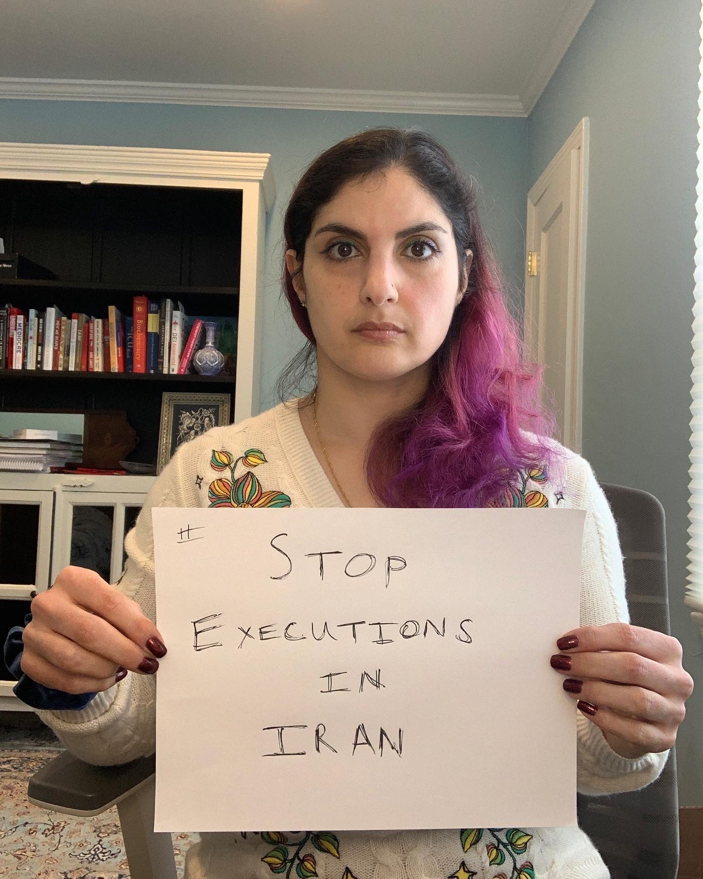 Selfie sitting in a chair, hair to the side, wearing a white floral sweater. Partially empty bookshelf behind me. Holding a sign that says #StopExecutionsInIran