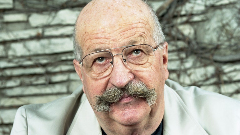 Gene Wolfe sitting in front of a stone wall with brown vines on it