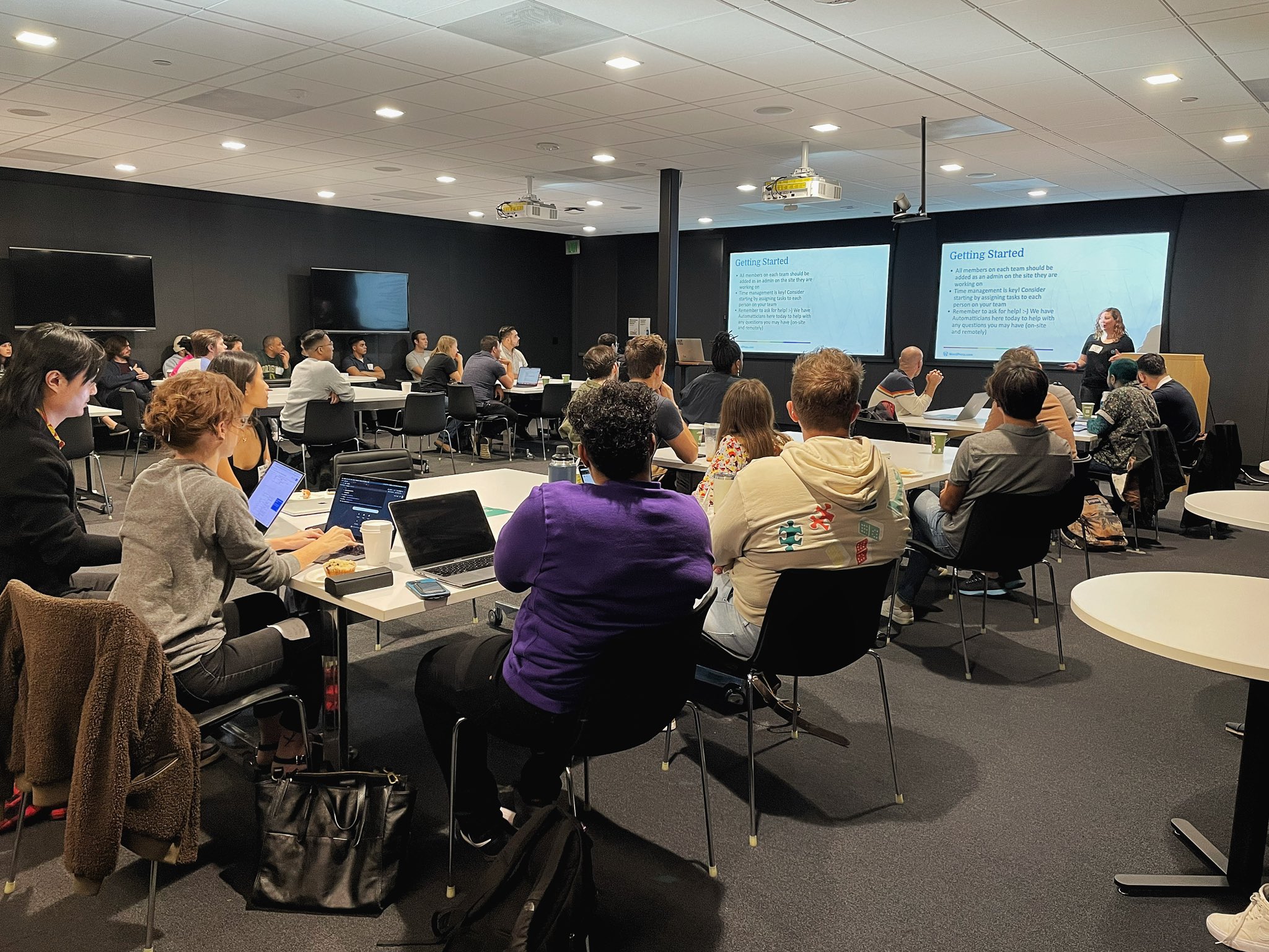 A Happiness Engineer from WordPress.com is giving a presentation to a room full of volunteers during the Out in Tech Digital Corps Hackathon