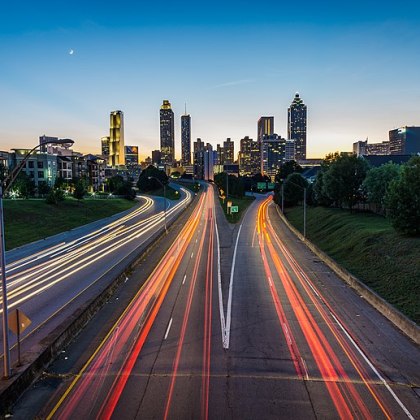 Long exposure of highway with downtown skyscraper in the background of an unidentified city. There are green grass hills on both sides and a bright moon in the sky.