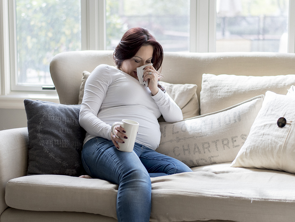 pregnant woman lying on a sofa and blowing her nose while holding a cup in her right hand