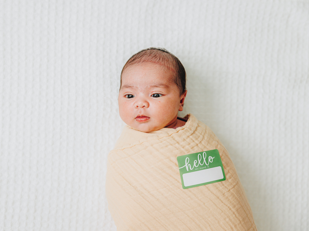 A newborn baby wrapped in a swaddle with a Hello My name is sticker. 