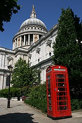 White stone building with tower topped with a dome. In the foreground are trees and a red rectangular vertical box with windows.