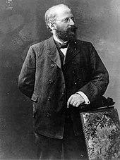 Eduard Bernstein standing next to a chair and looking rightwards. He is resting his hand on the chair.