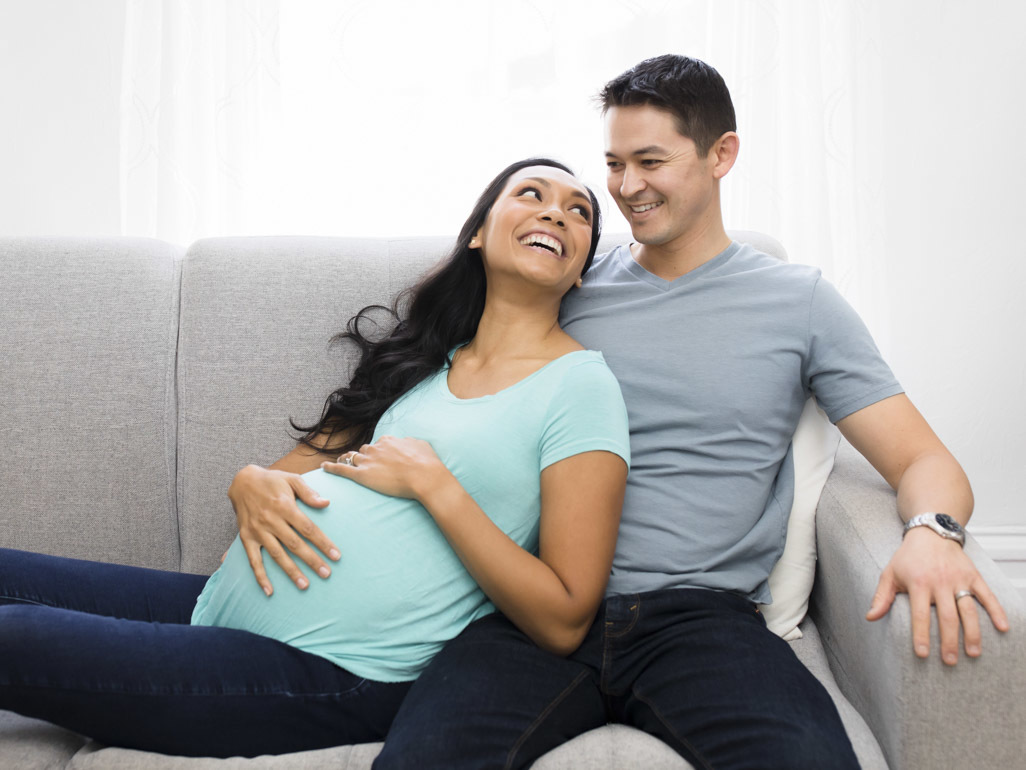 woman sitting on a sofa next to her partner, resting hand on her pregnant belly to feel her baby’s movements