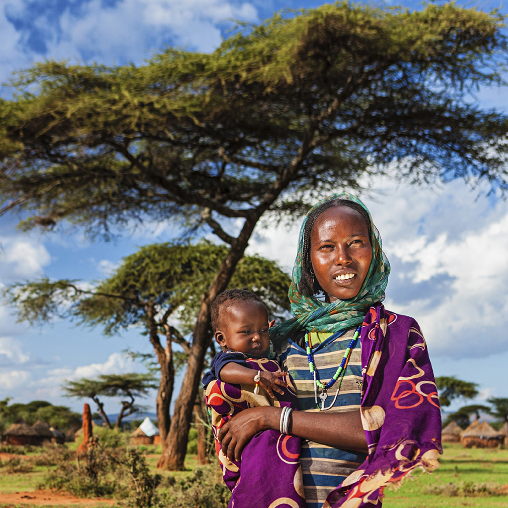 woman wearing head wrap holding baby in arms in African village