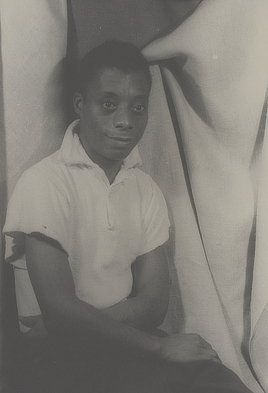 James Baldwin sits for a photographic portrait in front of a draped backdrop.