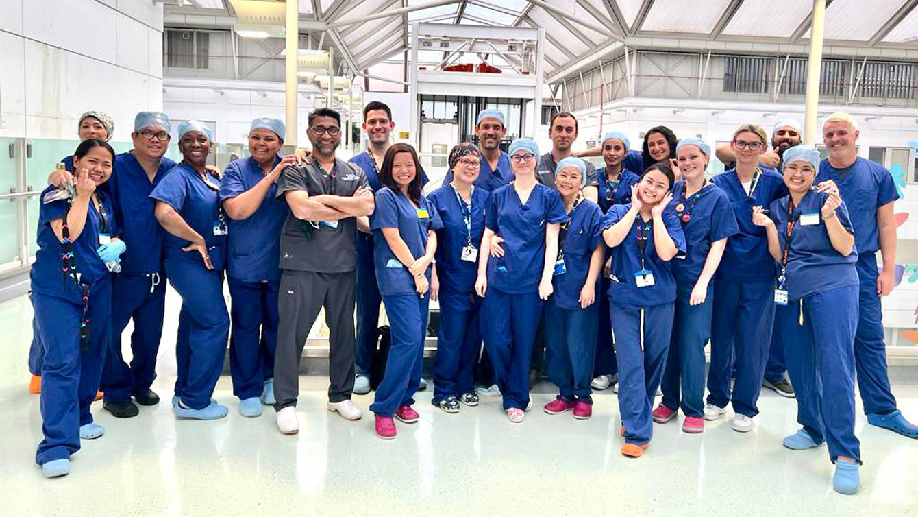 21 members of the gynaecological surgical team at Chelsea and Westminster Hospital stood in an open indoor space. All members of staff are wearing their surgical scrubs and are smiling into the camera. 