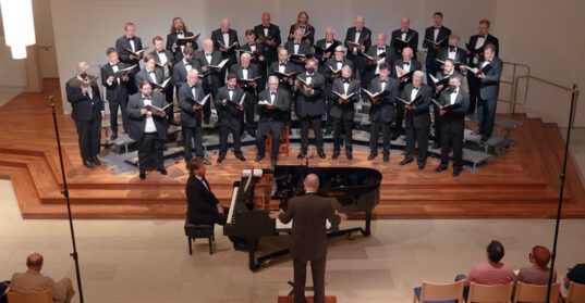 A group of men in formal attire on a raised platform, singing while accompanied on piano and under the direction of a conductor