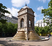 Farbige Eckansicht eines Brunnens im Renaissance-Stil mit zwei Treppen, über die Wasser fließt. Über den Stufen ist ein quadratischer Pavillon mit vier Rundbögen und Figurenreliefs, die von Giebelfeldern überdacht sind. In der Mitte ist eine graue Kuppel. Im Hintergrund des Platzes sind Bäume und Häuser.