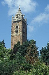 An ornate brick tower surrounded by trees. The tower has balconies and is surmounted by a pitched roof with an ornate figure at the apex.