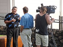 A man in a blue shirt, in front of a woman manning a camera, both engaging with a man in a black and blue shirt.