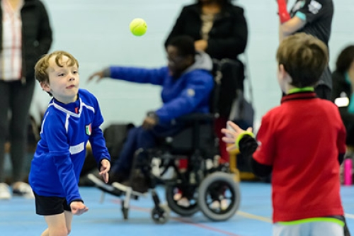 A child throws a tennis ball to another child. There is a wheelchair user in the background.