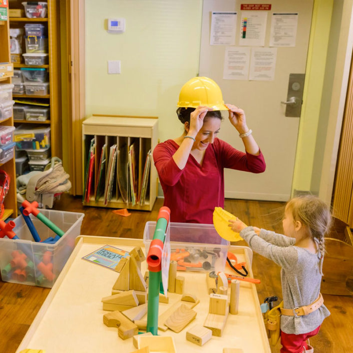 A toddler and a person are creating a human face out of child art at a table in a library.