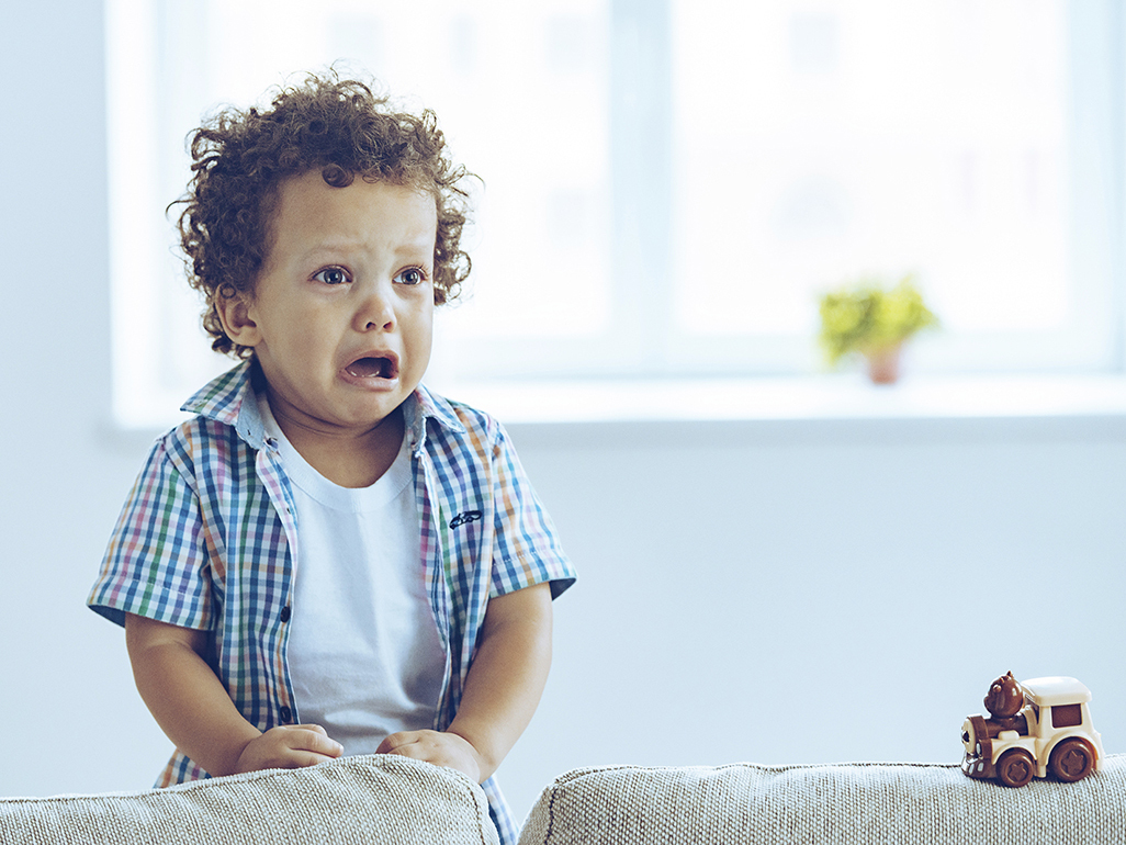 Little boy standing behind a sofa, looking upset