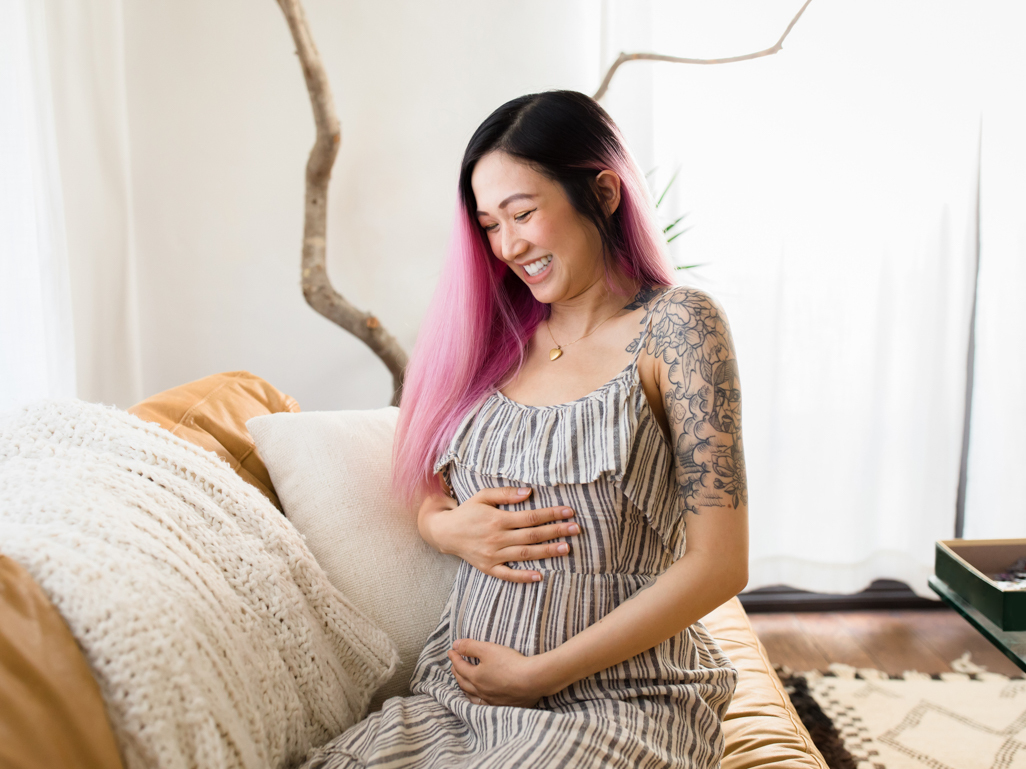 Mulher grávida branca de cabelo rosa sorri, segurando a barriga