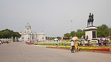 A large plaza with a bronze statue of a man riding on horseback; beyond the plaza is a large two-storey building with a domed roof, arched windows and columns