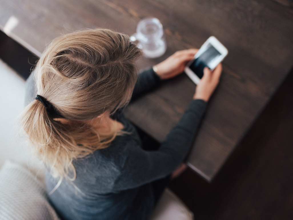 An aerial view of a woman sitting at a desk, looking at her phone.