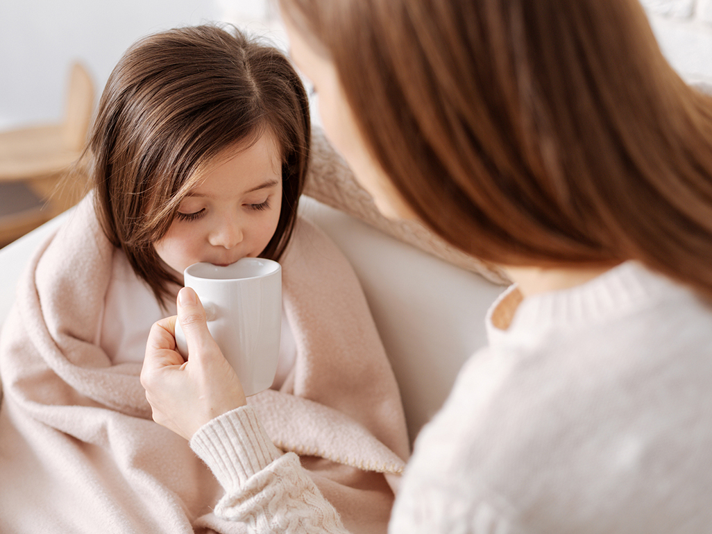 mother holding a mug while a seemingly under the weather child drinks from it