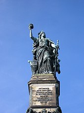 Monument of robed woman, standing, holding a crown in one hand and a partly sheathed sword in another