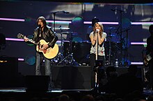 A long shot of a female performer, with long brown hair, singing into a microphone. Both of her hands rest on a microphone stand decorated with jewels. To her right is a male musician wearing a leather jacket, playing an acoustic guitar and also singing into a microphone.