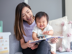 Woman sat with laughing baby on lap reading a book