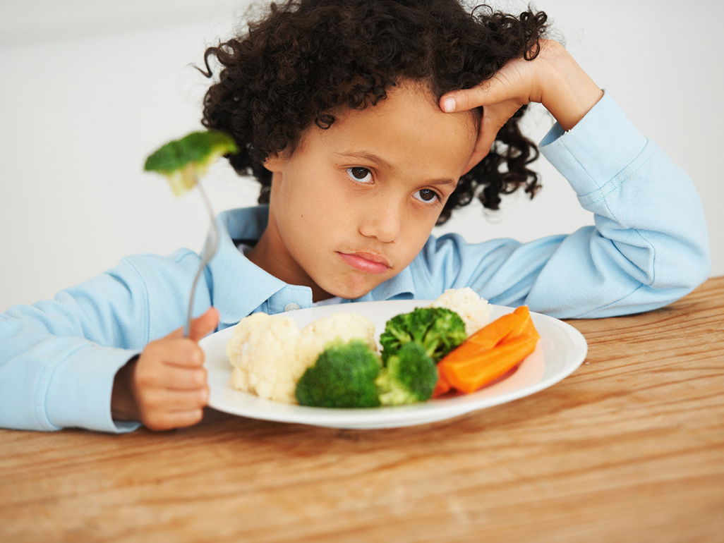 curly child holding a fork with broccoli and looking sad, while there are more vegetables in his plate