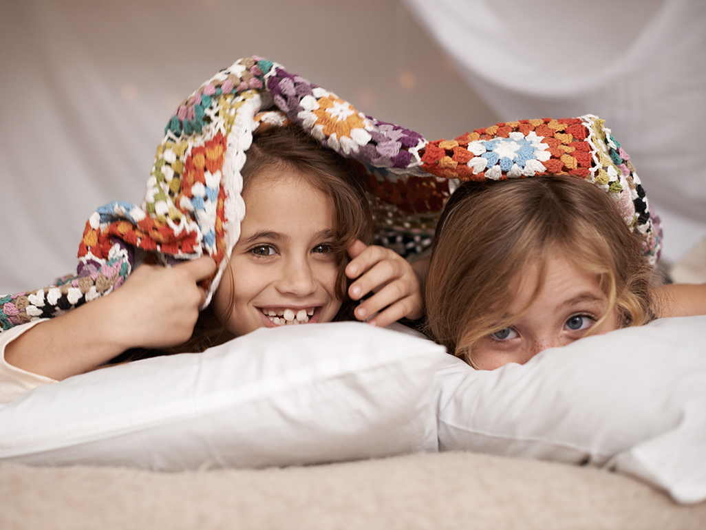 two girls lying on bed with blanket over head, only the faces peeping out