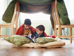 two kids reading a book on pillows, under a blanket