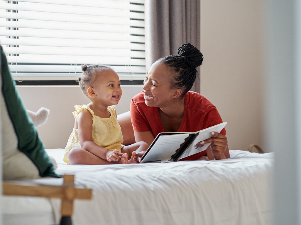 Toddler on bed reading book with mother 