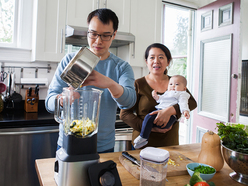 Mother holds baby while dad tips vegetables into a food maker