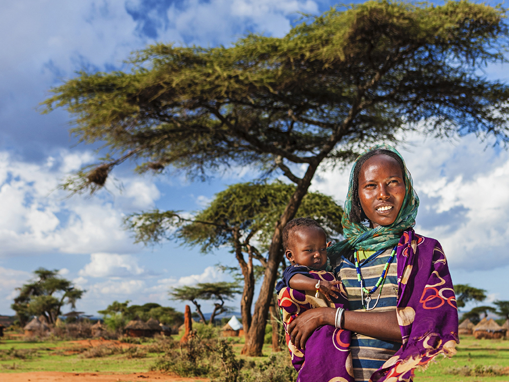 woman wearing head wrap holding baby in arms in African village