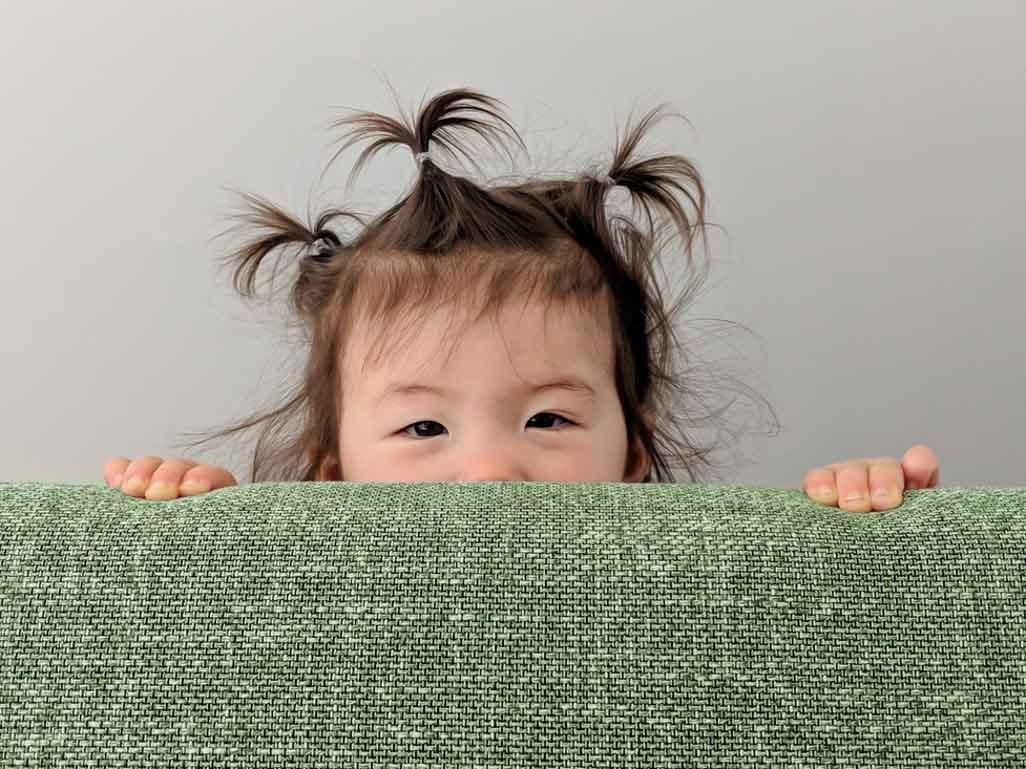 A toddler peeking their face out from behind a couch