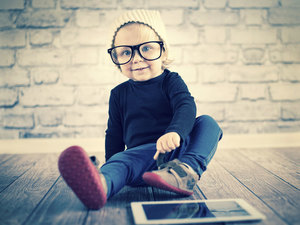 A toddler wearing a beanie and glasses sitting in front of a tablet