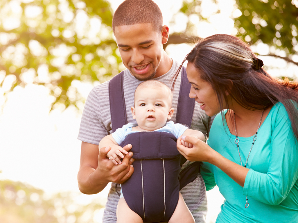 parents walking outside, father carrying their baby in a carrier, while mother is holding baby's hand