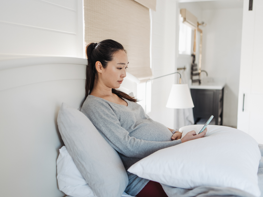 A pregnant woman lying on a bed and looking at a cell phone 