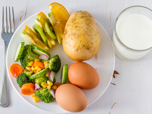 plate with potato, hardboiled eggs, kiwi, orange, broccoli, carrots, and corn, plus a glass of milk