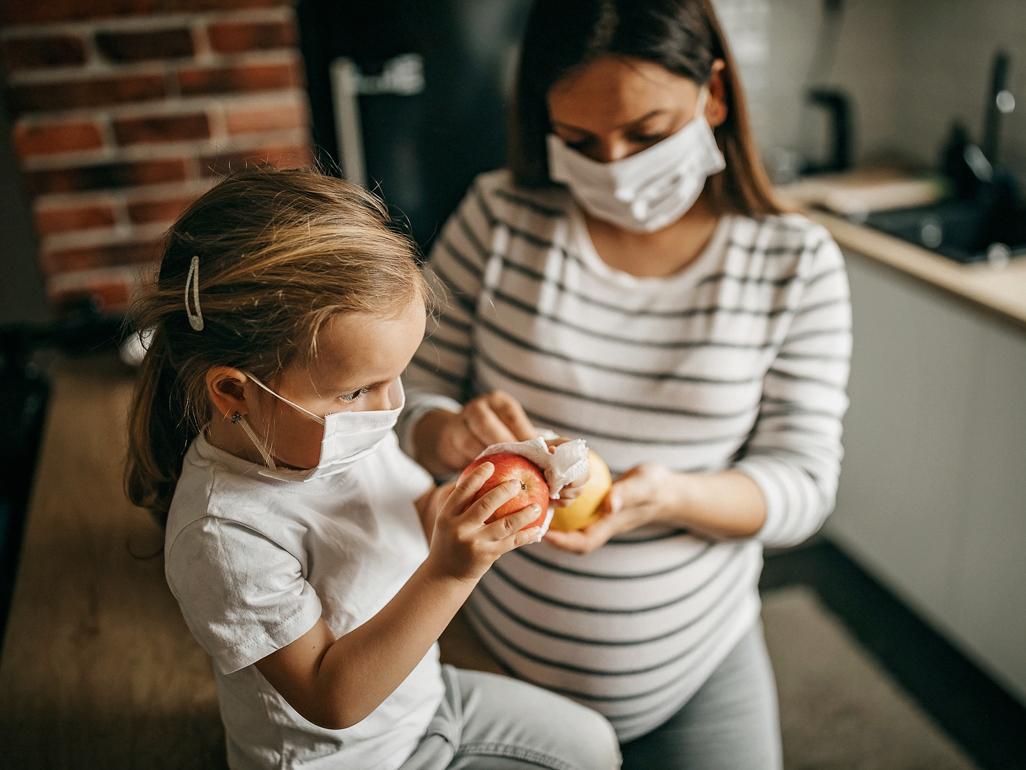 A mom and her daughter, both wearing face masks, cleaning fruit in a kitchen.