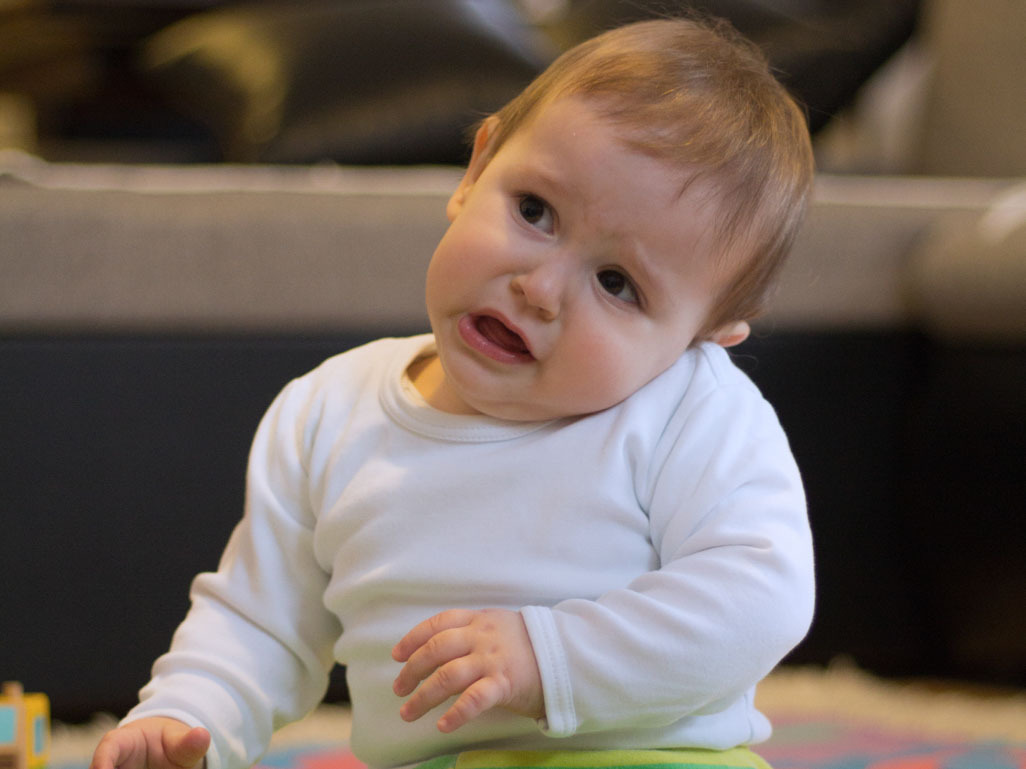 A baby sits on the floor, holding their ear to their shoulder, text reads: BabyCenter Answers, Ear infections in babies