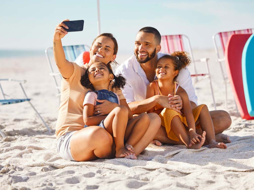 family of mom, dad and two young girls taking a selfie on a beach