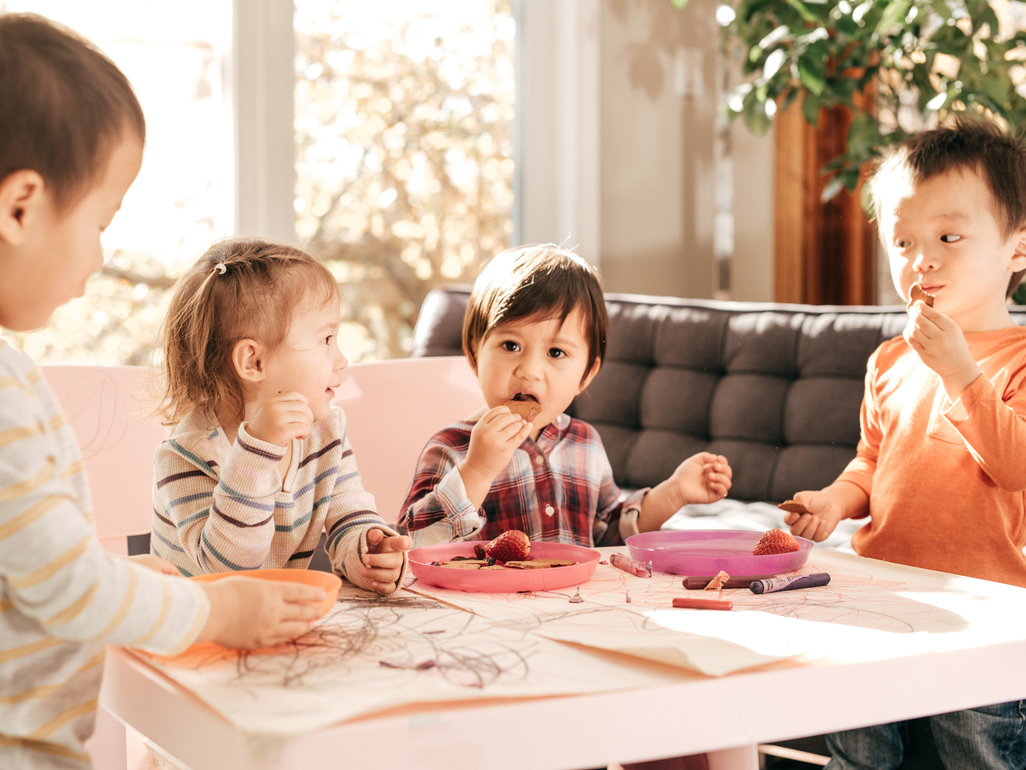 three boys and a girl sitting at a table, eating crackers and fruit