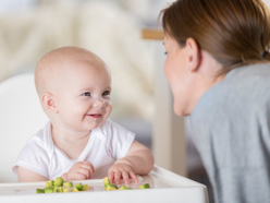 Six month old baby in highchair eating first foods while mum watches