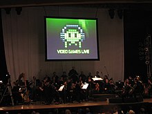 A photograph of an orchestra on a dimly lit stage. Above the group is a projection screen with a black, white, and green image of pixel art. The pixel art is an oval object wearing headphones with eyes and four tentacles. Below the pixel art is the phrase "Video Games Live".