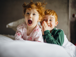 A toddler girl lying on a bed screaming; her brother is smiling in the background.