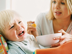 Boy in highchair refusing breakfast from his mother