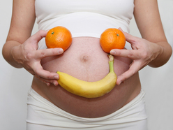 Mom holding a banana and two oranges up to her pregnant belly, making a smiley face around her navel.